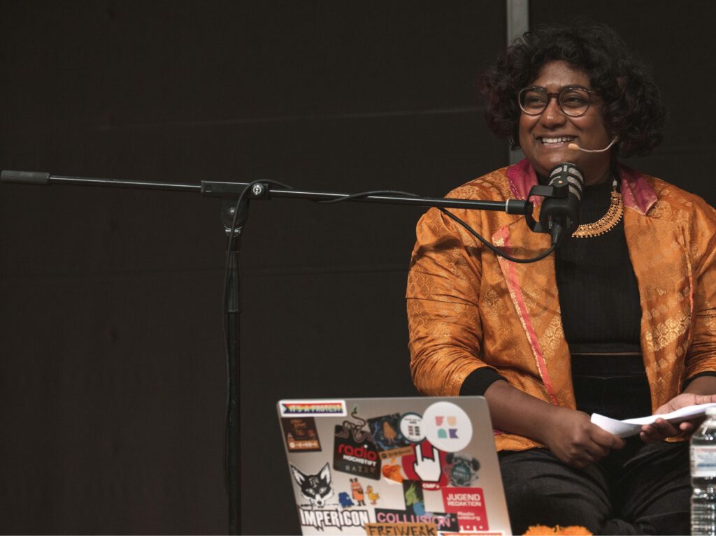 Abby in einem Blazer aus Saree-Stoff. Schwarzer Hintergrund. Abby sitzt hinter einem Mikrofon und lacht.